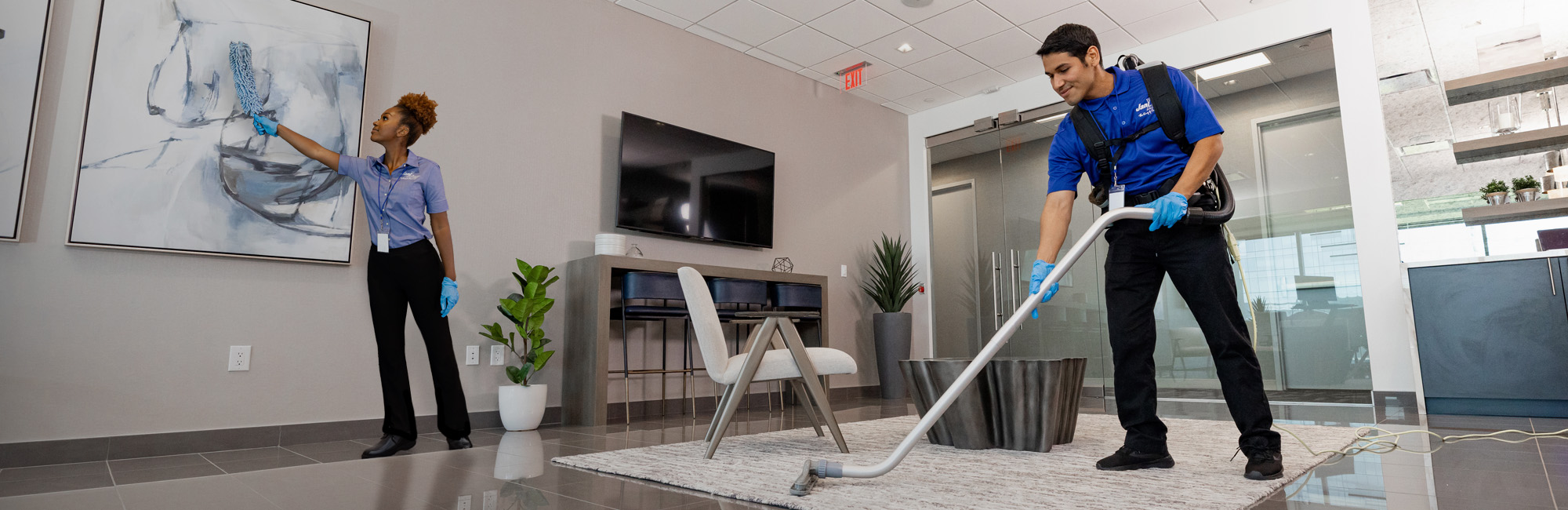 Two workers in a modern office space, representing Jani-King office cleaning services, tidy a room. One vacuums the carpet while the other wipes a framed picture. They wear blue shirts and gloves, with cleaning equipment visible amid the TV, chairs, and minimalist decor.