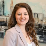 A woman with long, wavy brown hair is smiling. She is wearing a light-colored blazer over a white top.